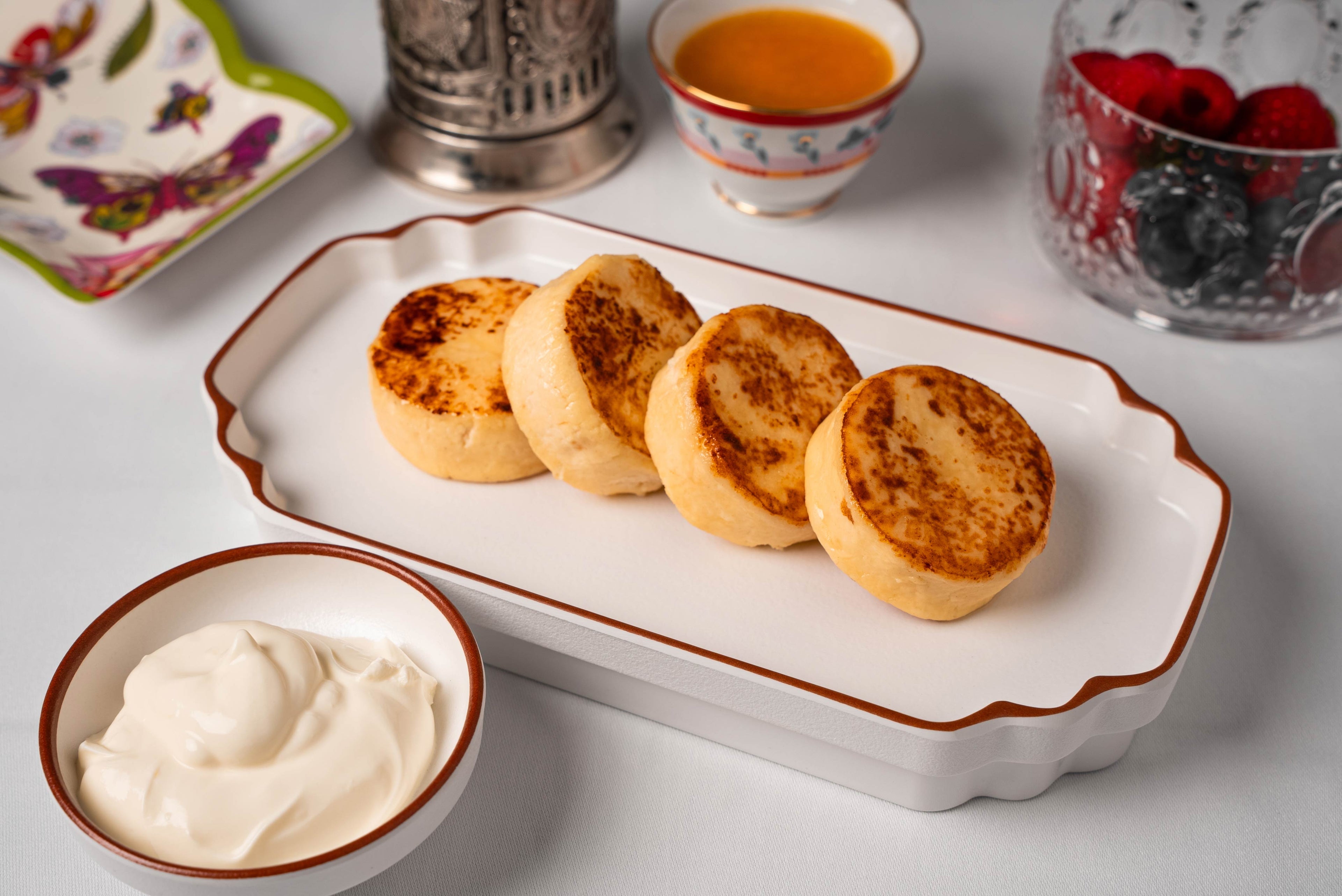 A white Cosmopolitan melamine tray with terracotta rim holding golden treats, set beside a butterfly dish and a glass bowl of fresh berries