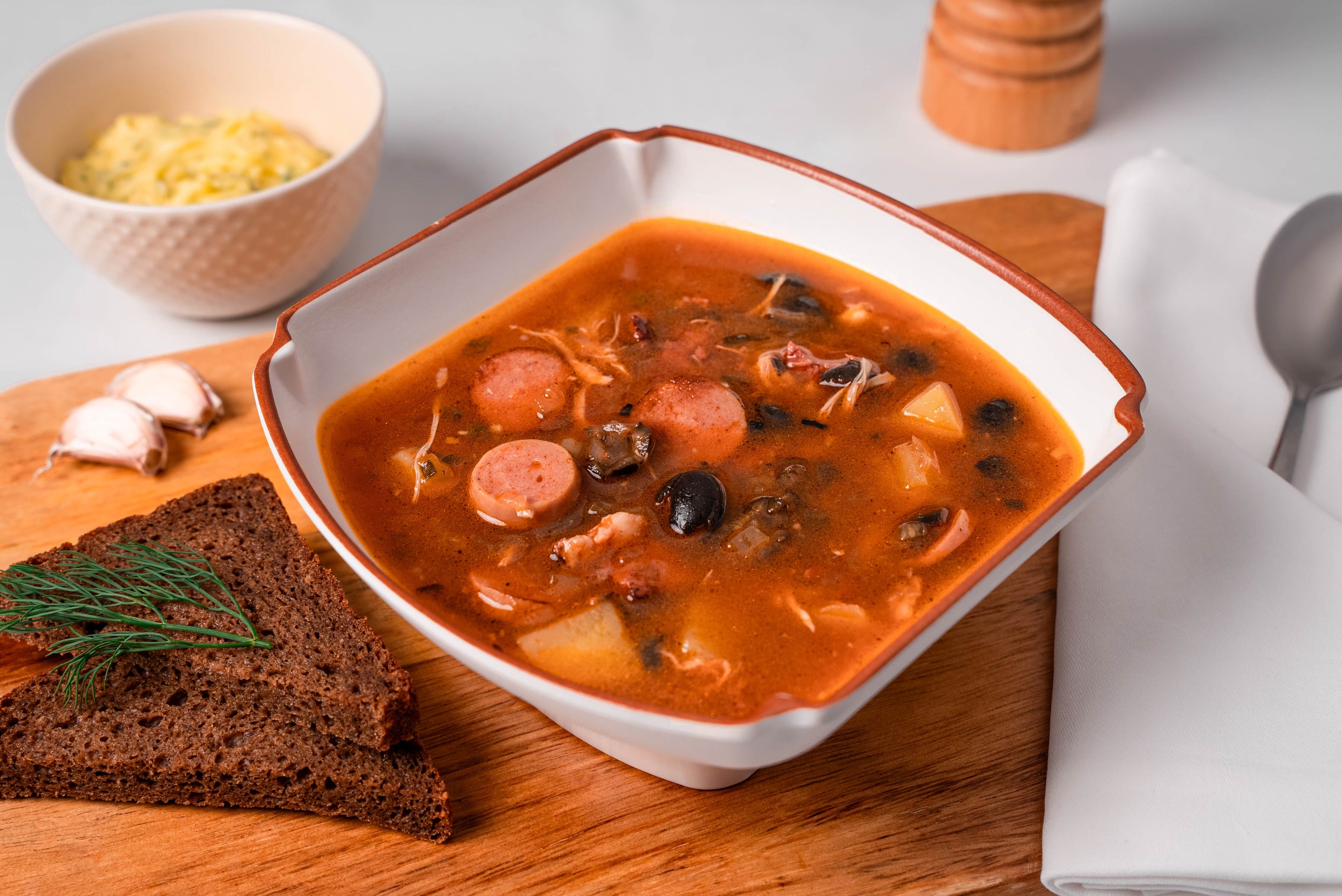 Hearty soup served in a square white Cosmopolitan melamine bowl with a terracotta rim on a wood board, with rye bread alongside