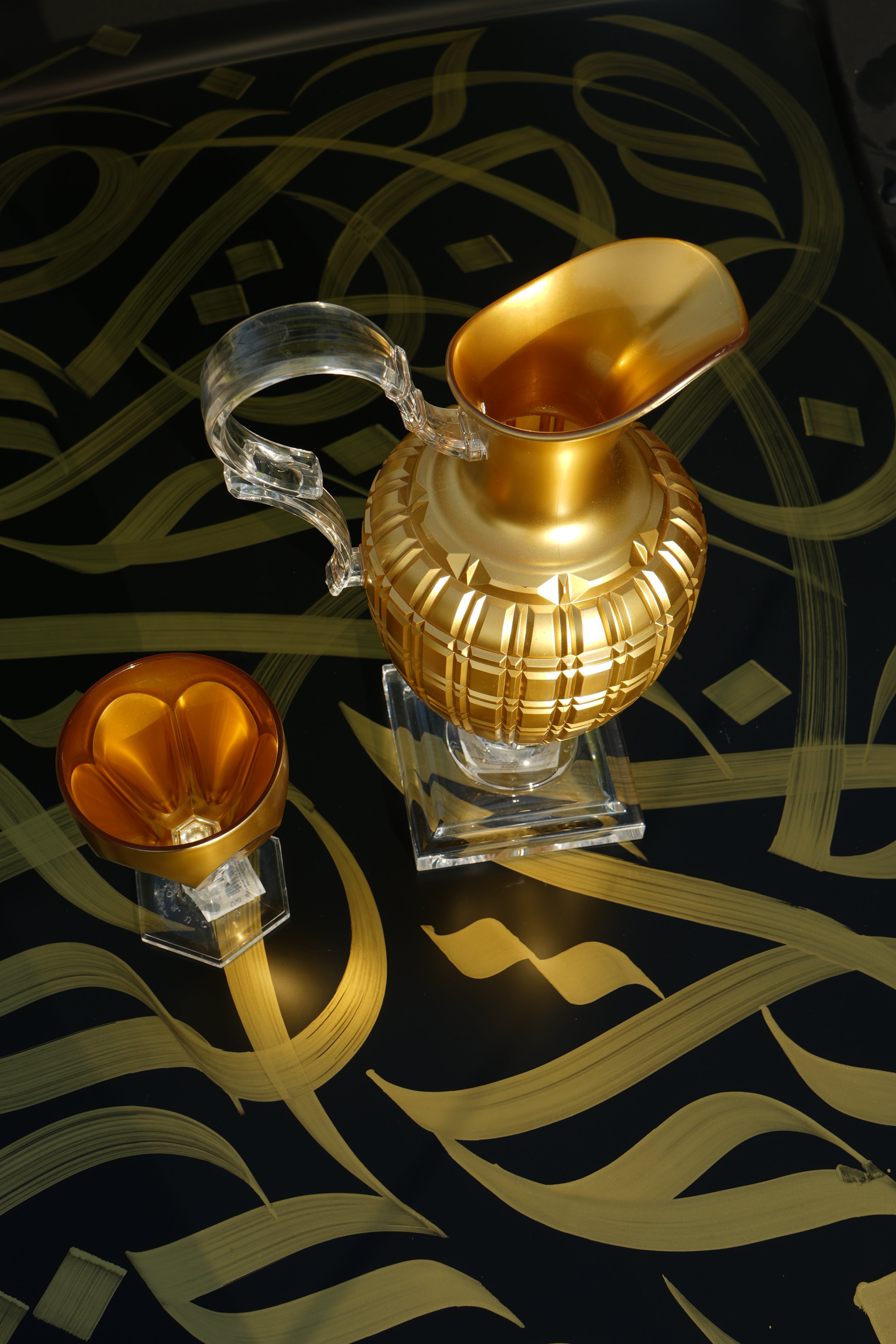 Gold Mario Luca Giusti pitcher and amber goblet on a black Arabic-calligraphy surface, evoking a warm Bahraini majlis welcome