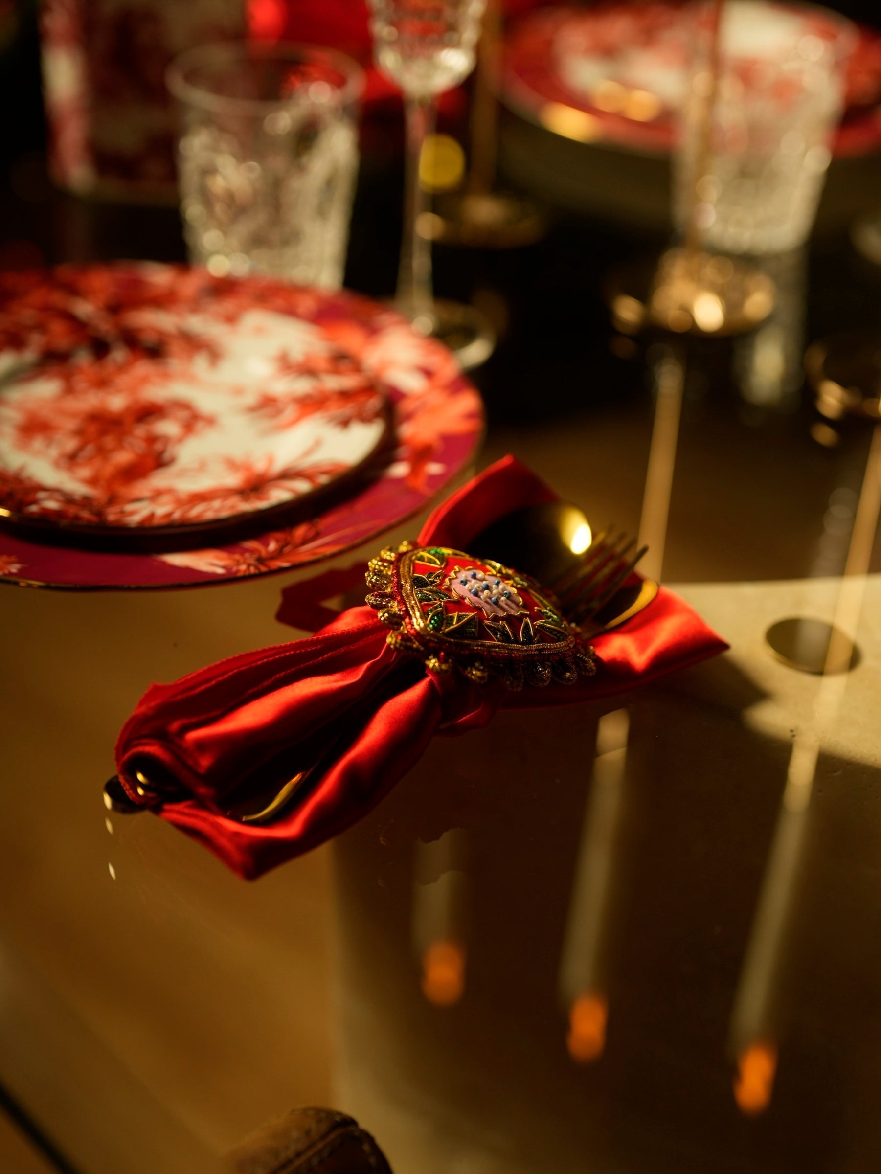 Red satin napkin tied with an ornate jewelled napkin ring, gold flatware and a red-and-cream patterned Italian porcelain plate set under warm candlelight.
