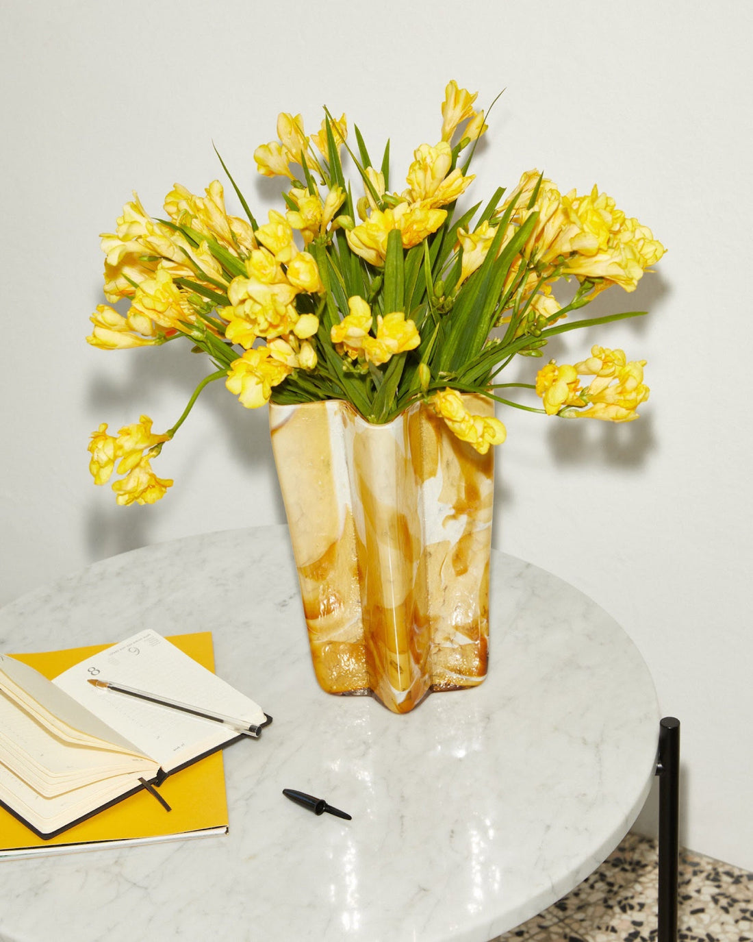Amber and white swirled glass vase holding yellow freesias on a marble side table