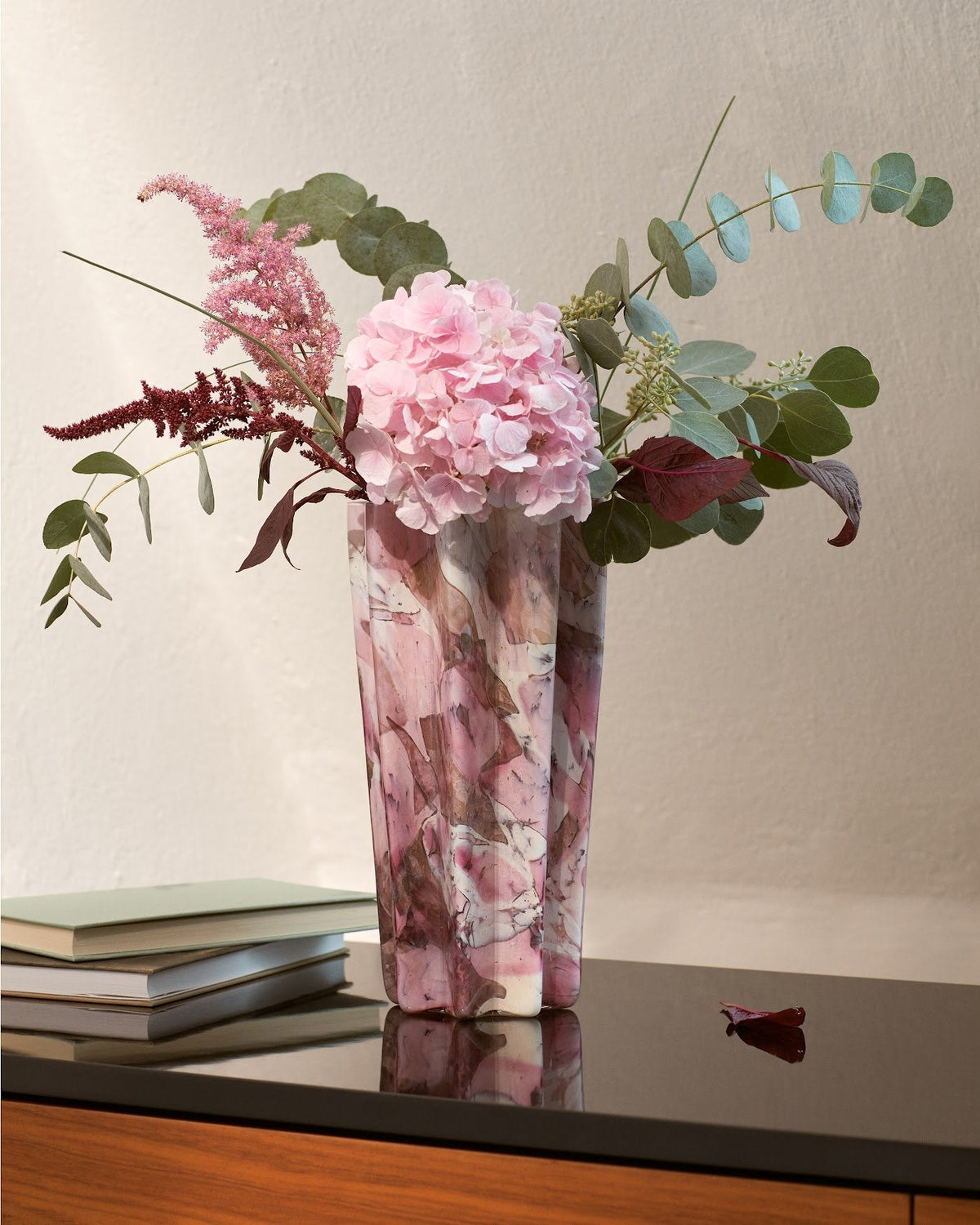 Pink Murano glass vase with hydrangeas beside stacked books in a warm winter interior