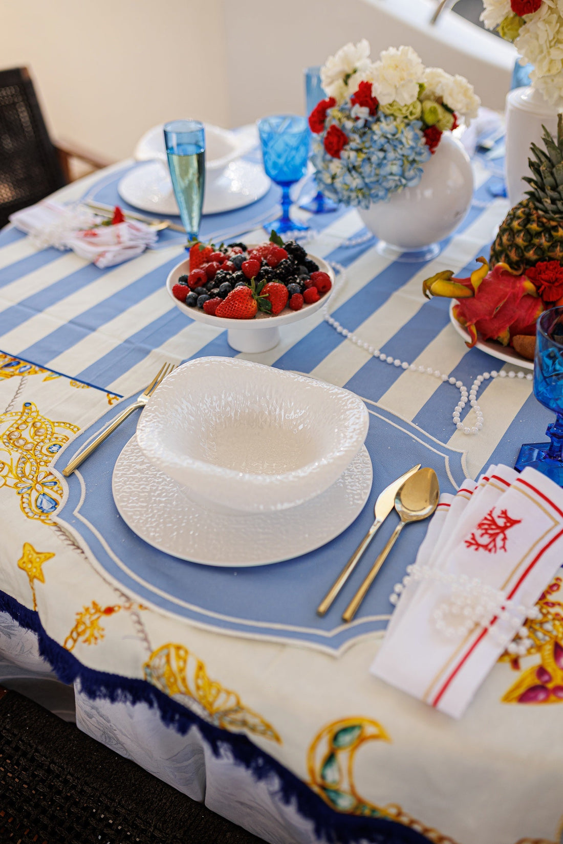 Table setting with blue placemats, white textured plates, gold cutlery and a berry stand on striped linen