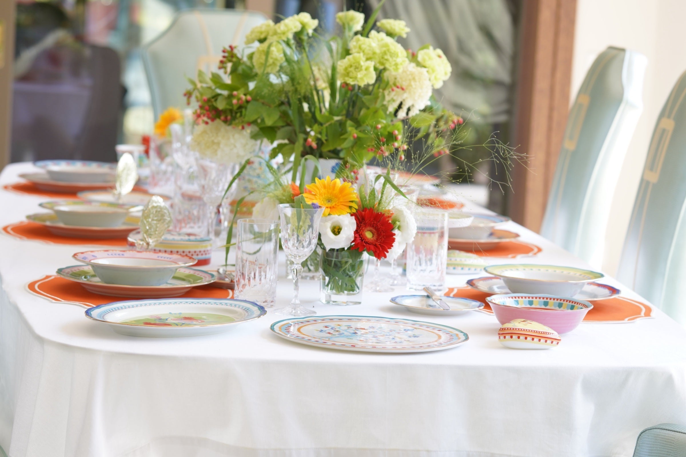 Colourful Italian tablescape with mixed-pattern porcelain, orange placemats, crystal glasses and a garden flower centrepiece