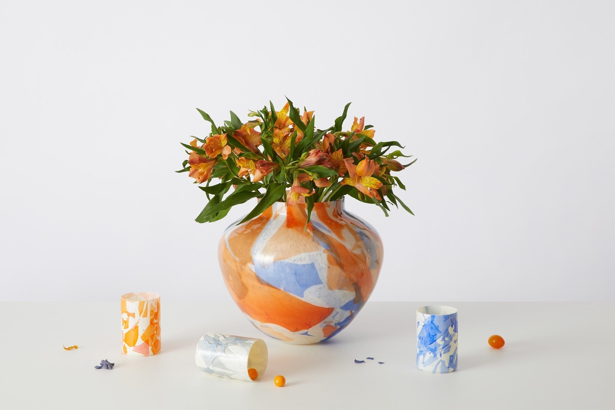 A Stories of Italy Nougat-technique Murano glass olla vase in fused orange, cobalt and white, beside two matching marbled tumblers on a clean white studio surface.