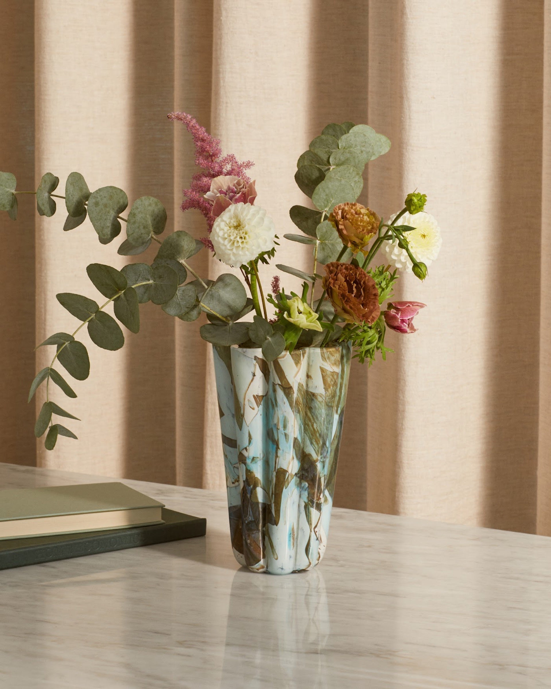 Marbled art-glass vase with dahlias and eucalyptus on a marble console in soft window light