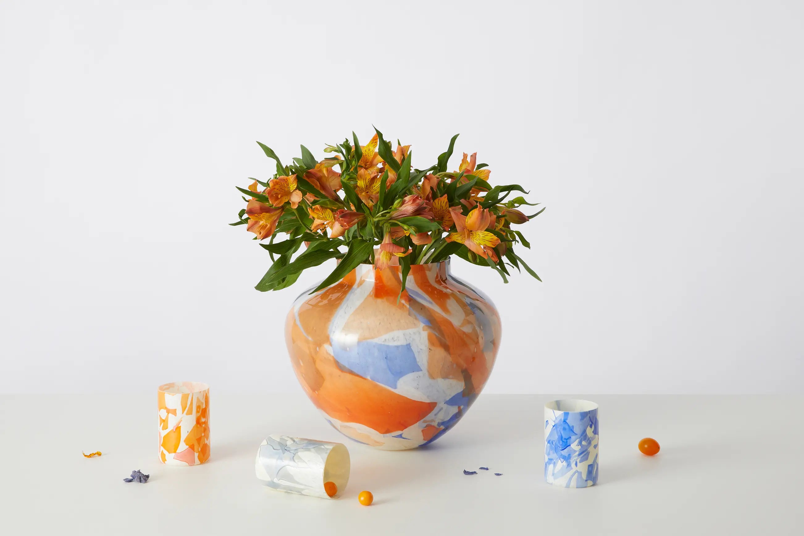 Stories of Italy Summer Olla vase in orange and cobalt Murano glass holding alstroemeria, with smaller Nougat tumblers arranged on a pale tabletop.