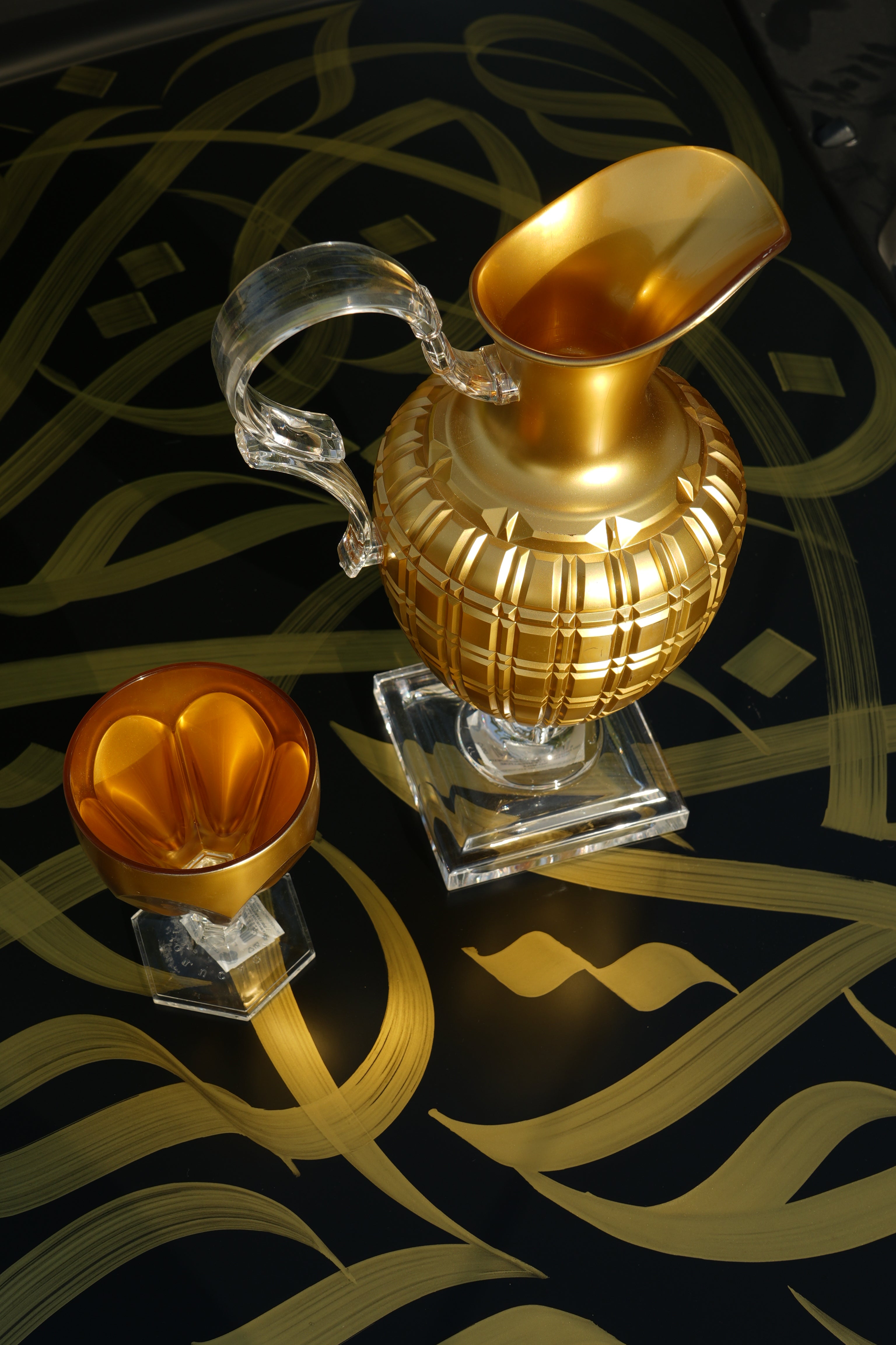 Gold Palla pitcher and amber Lente vase by Mario Luca Giusti arranged on a black surface with gold Arabic calligraphy patterns.