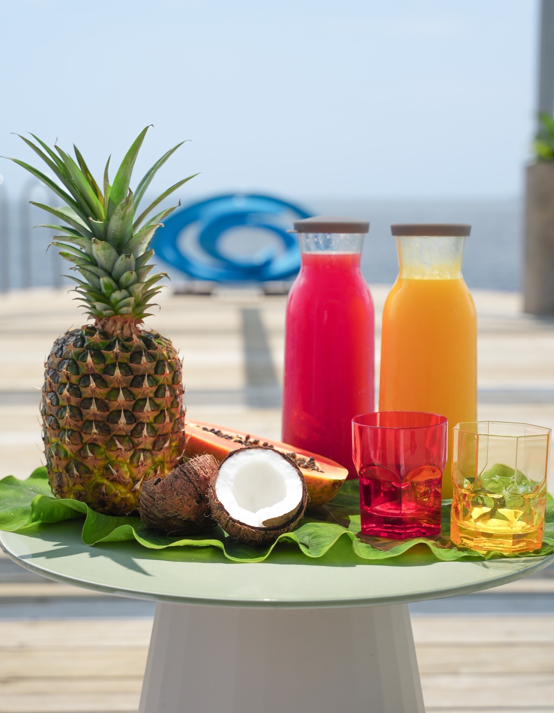 Translucent red and yellow acrylic tumblers with cold-pressed juice carafes beside tropical fruit on a cake stand at an outdoor table.