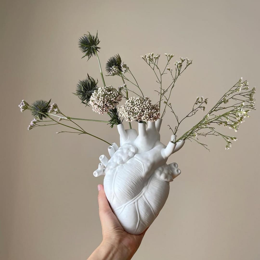 Love in Bloom porcelain vase shaped like an anatomical heart, held in hand with delicate flowers — Amprio Milano.