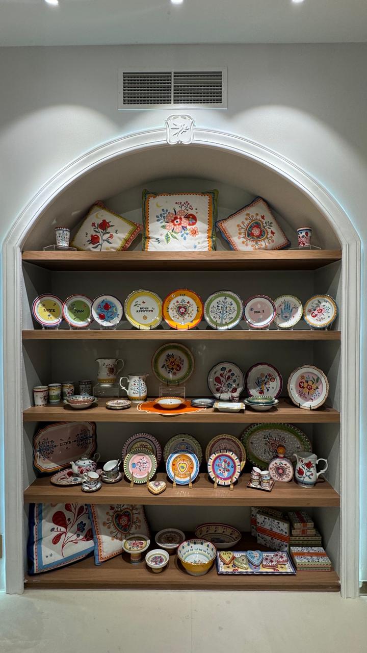 Mamma Mia showroom alcove with embroidered floral cushions, hand-painted Italian porcelain plates, jugs and teapots in joyful colours, and folded embroidered table textiles below.