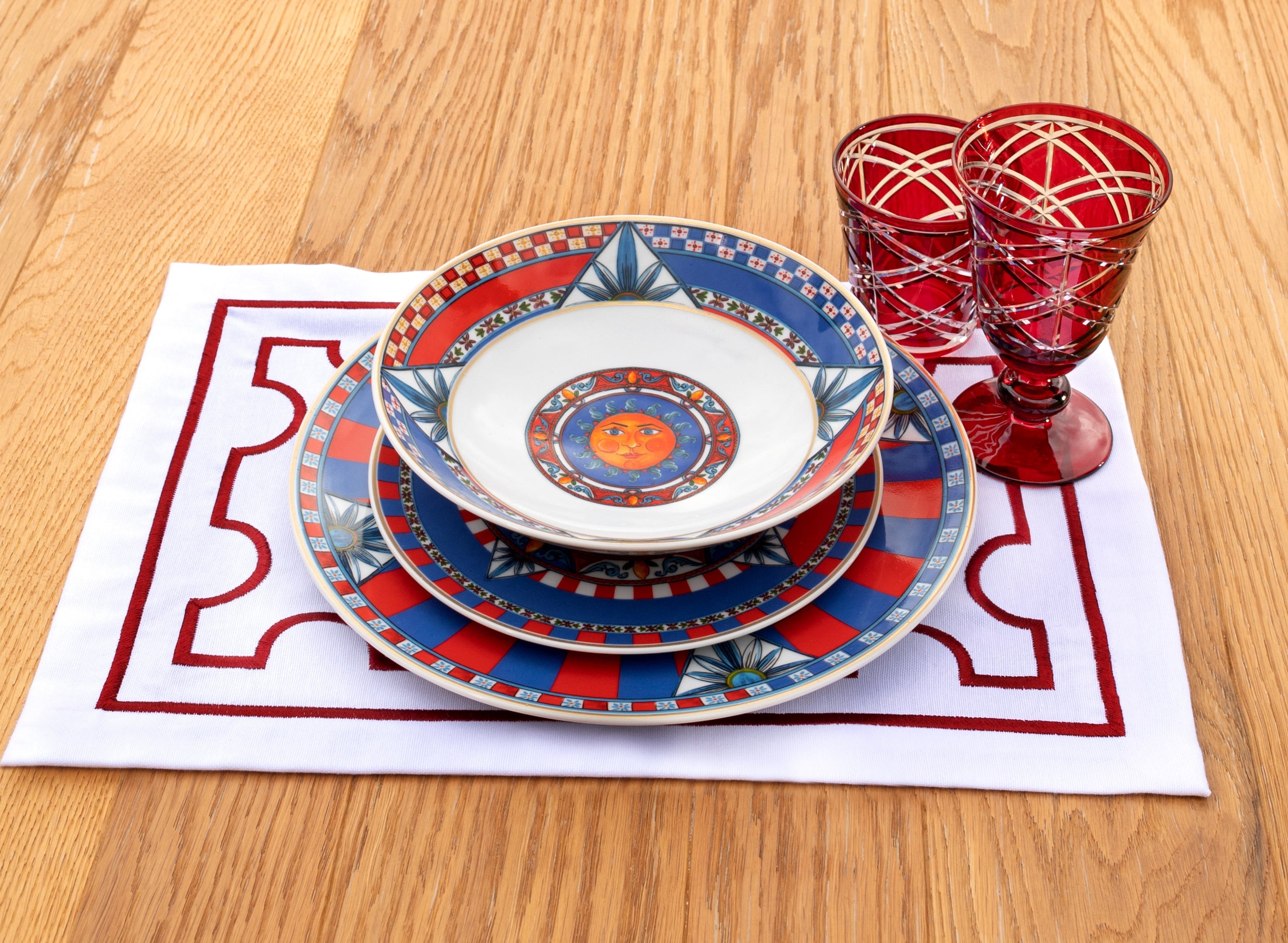 Sole Mio place setting — Sicilian sun motif plates with red patterned glasses on a wooden table by Amprio Milano.