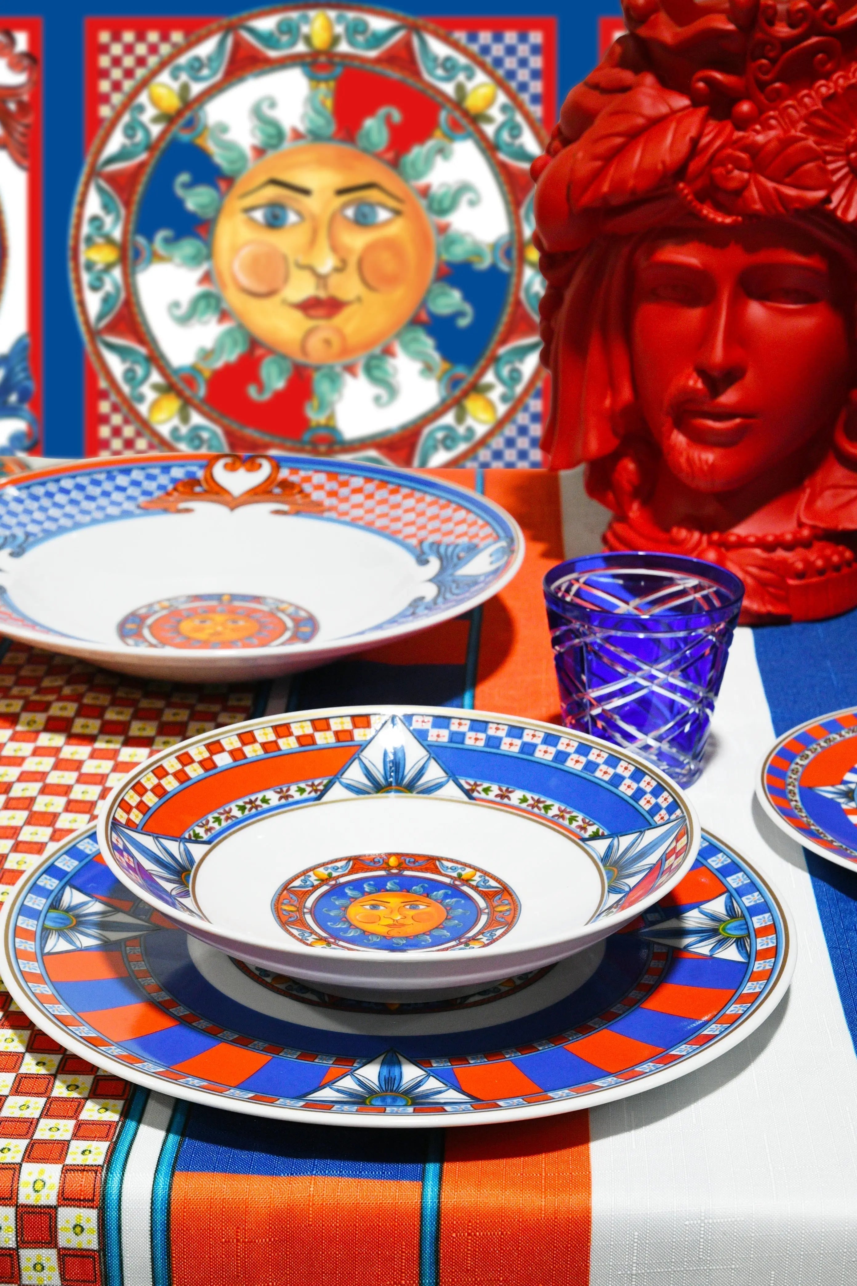 Sole Mio table setting — Sicilian sun motif plates in bold red, blue and orange with Moorish-head accent and coloured glass by Amprio Milano