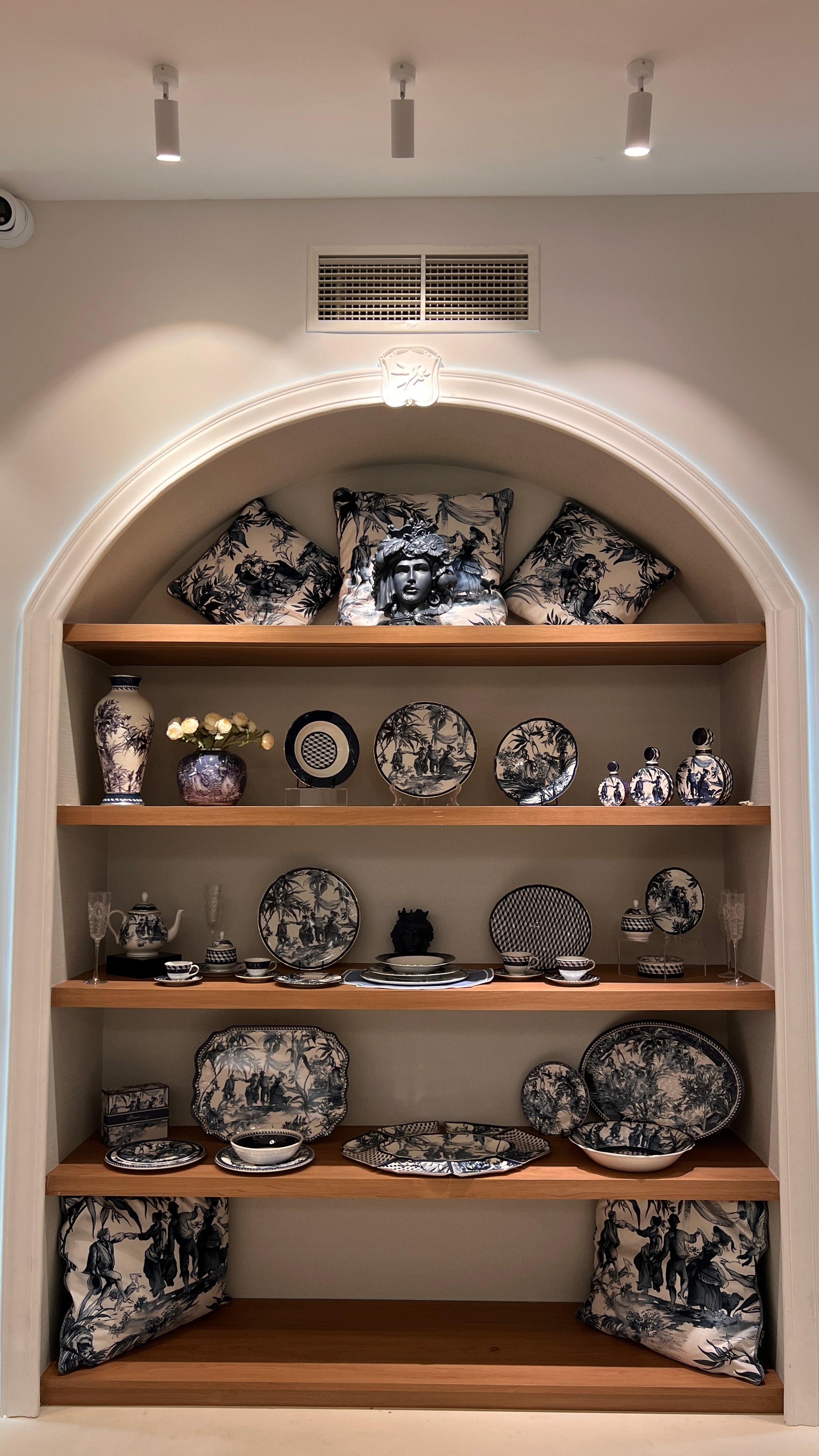 Calm blue-and-white Italian tableware display in a Muscat alcove — toile plates, vases and cushions arranged on warm wood shelves