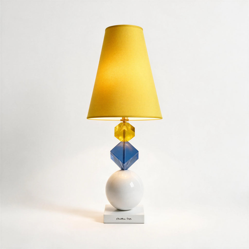 A modern table lamp with a yellow fabric cone shade, a faceted yellow crystal accent, a blue faceted crystal accent, a glossy white spherical base, and a white rectangular marble base, photographed on a clean white background.